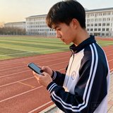 一位穿运动服的中国男大学生在校园操场边用手机查看球探体育比分，背景为大学田径场和教学楼